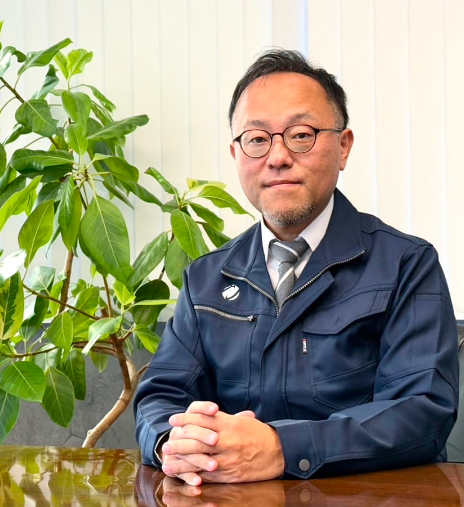 代表取締役：市岡 真二のバストアップ画像。テーブルの上で手を組んでいる。